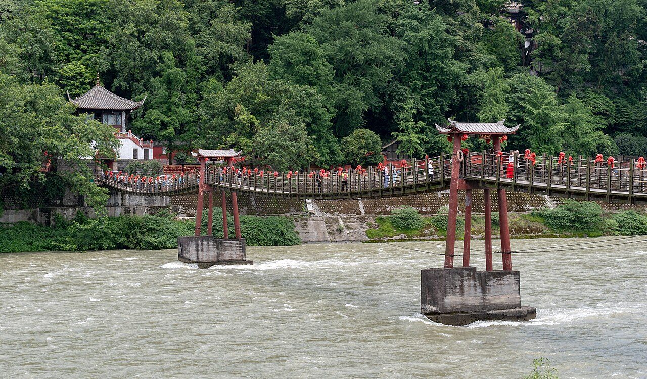 Die Hängebrücke Anlan über den Min-Fluss bei Dujiangyan, mit roten Holzpylonen und weißem Wasser darunter — Foto: xiquinhosilva / CC BY 2.0 über Wikimedia Commons