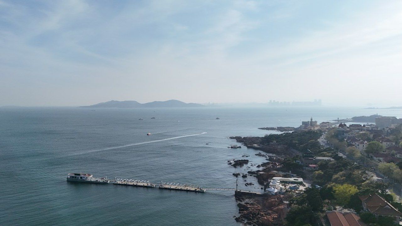 An aerial view of Qingdao's coastline — rocky headlands, a pier and the city skyline on the horizon; the far end of the Line 8 ride