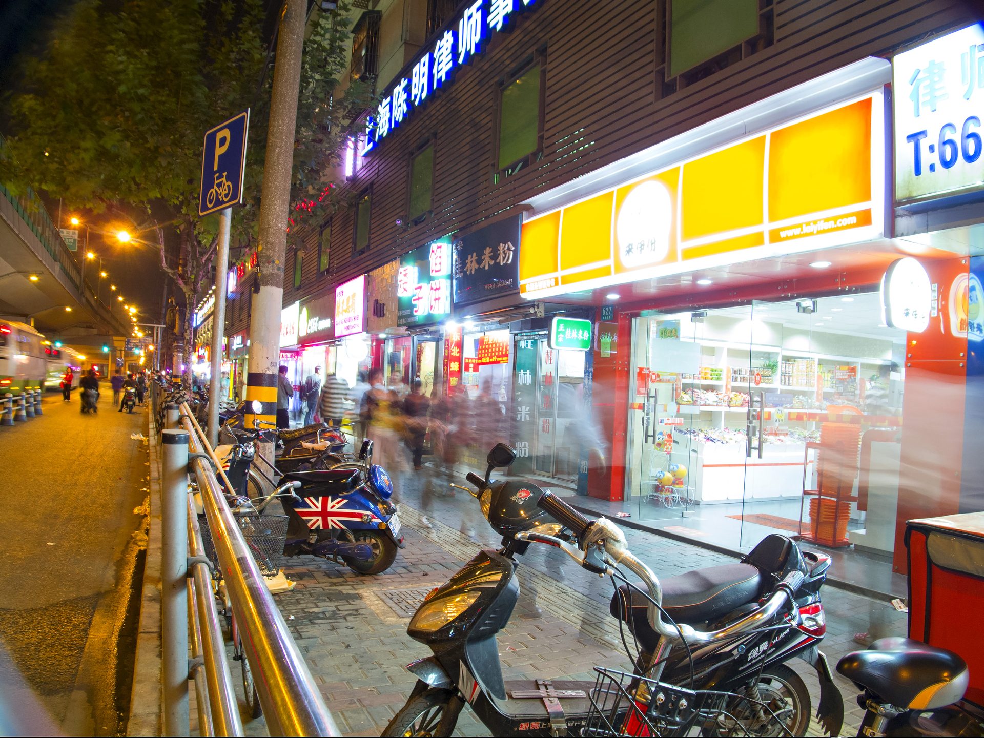 A Shanghai back-street food row at night, lit noodle shop and snack store signs — Photo: Михаил Лазарев / CC BY 2.0 via Wikimedia Commons